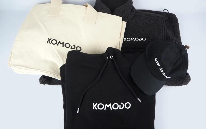 A flat lay of clothing items including a beige tote bag, a black hoodie, a black hat with 