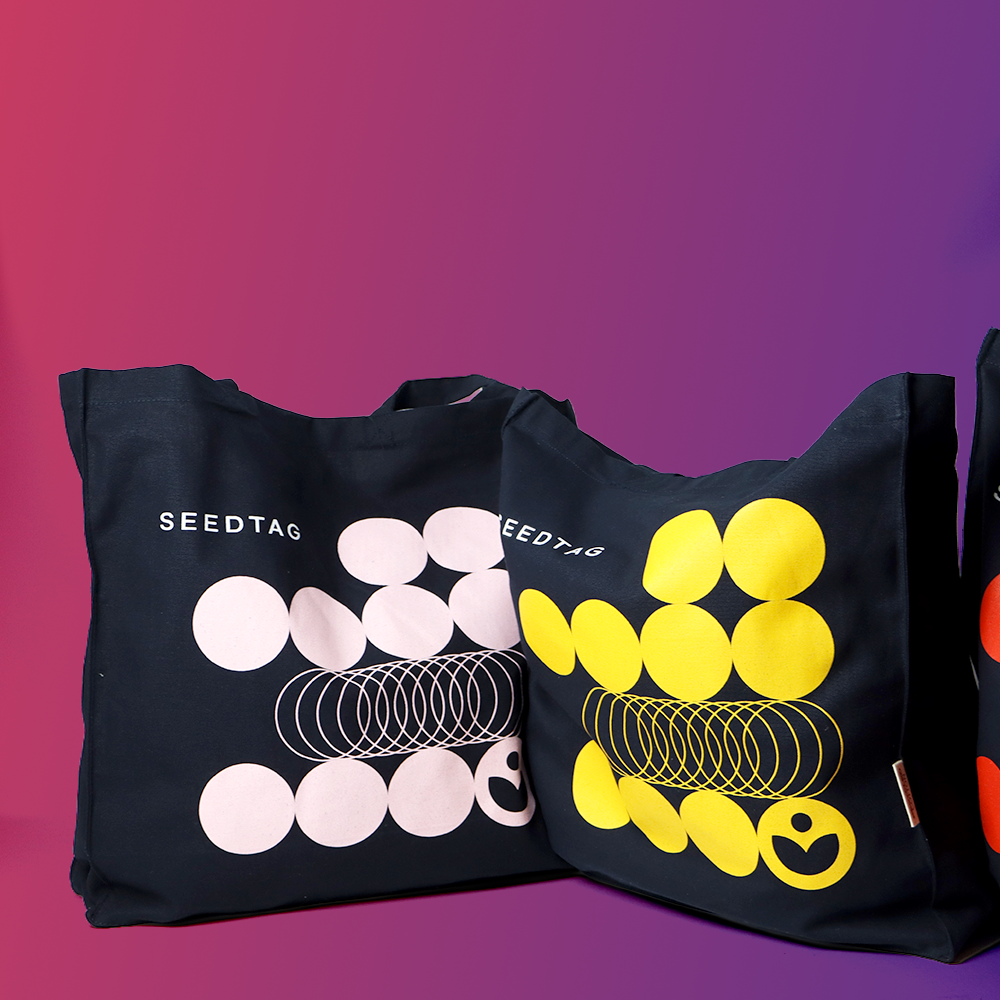 Three tote bags in black, featuring abstract designs in white, yellow, and pink, with the logo 