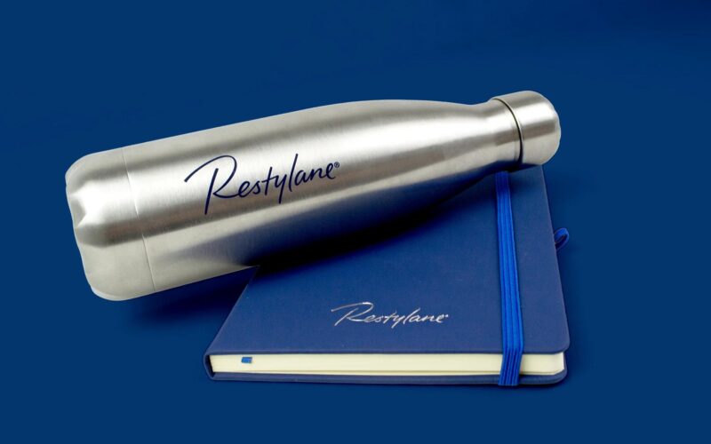 A stainless steel water bottle next to a blue notebook, both featuring the logo 