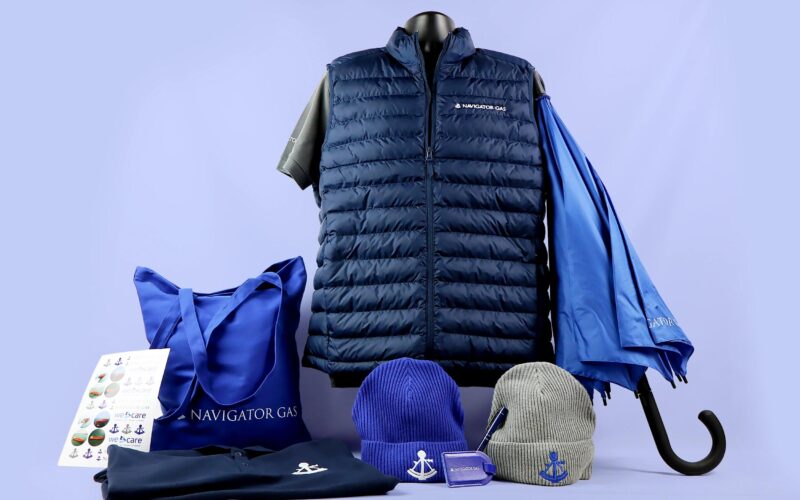 A collection of branded merchandise including a navy puffer vest, a blue umbrella, a blue tote bag, two beanies (one blue, one gray), a black polo shirt, and a sheet of stickers.