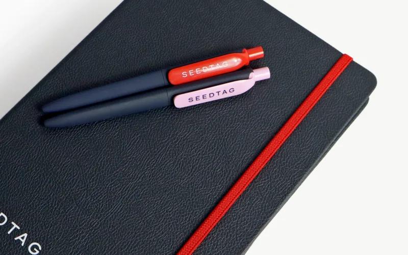 Two pens, one red and one pink, resting on a black notebook with a red elastic band. The notebook has 