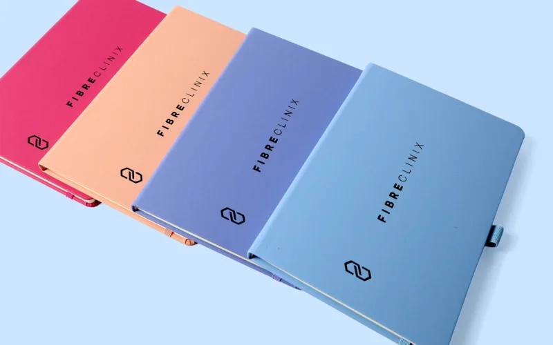 Four colorful notebooks from the brand Fibreclinix, arranged in a row on a light blue background.