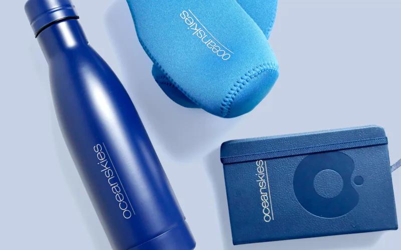 A blue stainless steel water bottle, a blue neoprene item, and a blue wallet or pouch, all featuring the brand name 