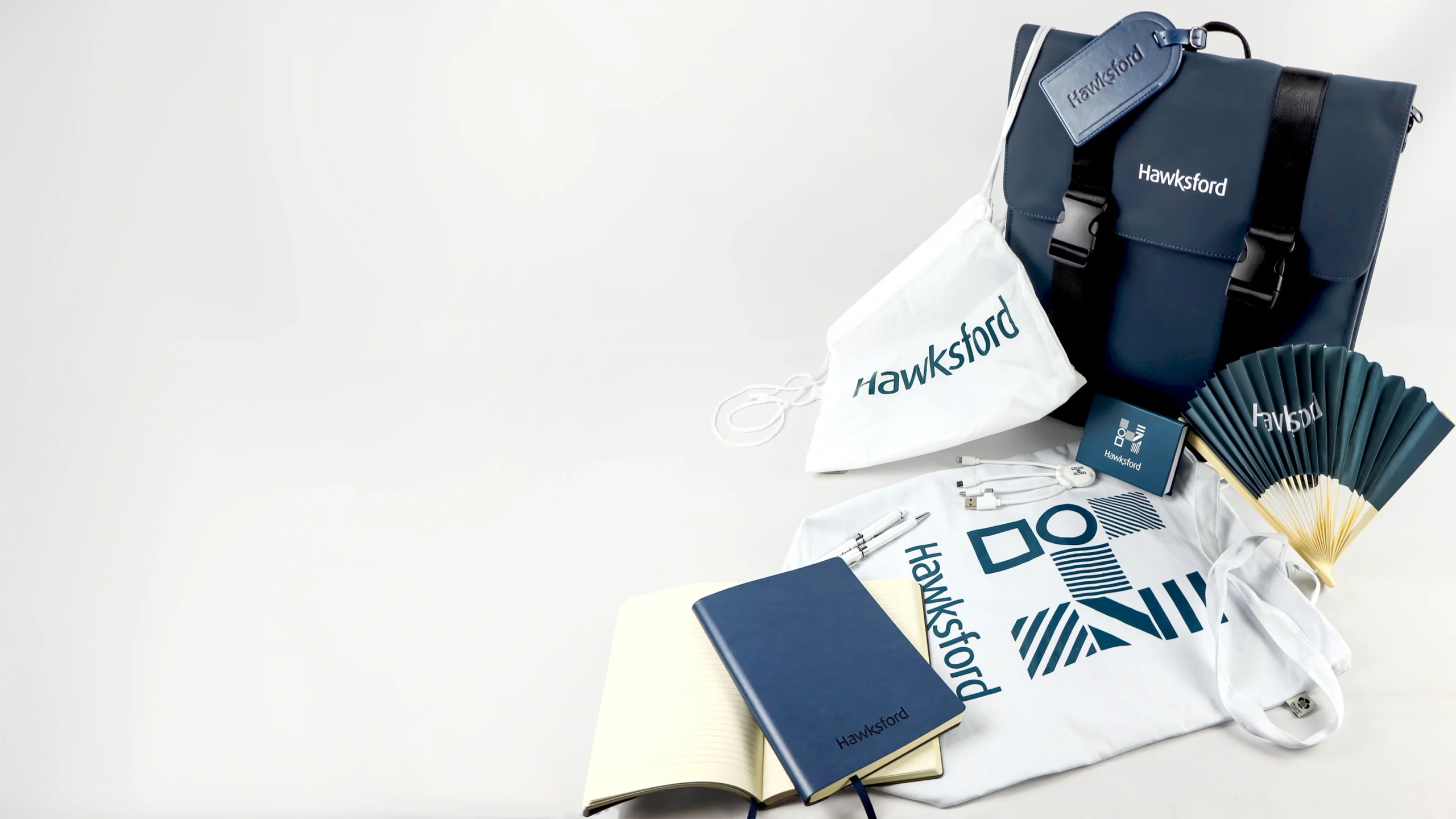 A flat lay of promotional items including a dark bag, a white tote bag, a blue notebook, a white t-shirt, a fan, and accessories like a pen and charging cables, all branded with the name 