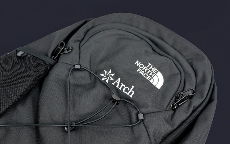 A black backpack featuring logos from The North Face and Arch, with adjustable straps and a mesh pocket.