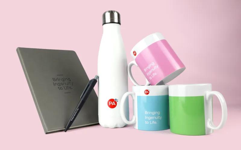 A white water bottle, three mugs in pink, light blue, and green, a gray notebook, and a black pen, all set against a pink background.