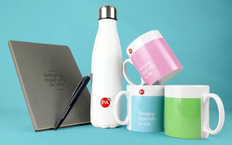 A collection of branded items displayed on a blue background, including a white water bottle, three mugs in pink, blue, and green, a gray notebook with the text 