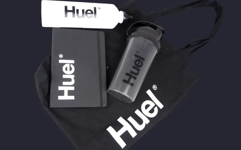 A black tote bag, a black notebook, a gray shaker bottle, and a white bottle, all branded with the Huel logo.