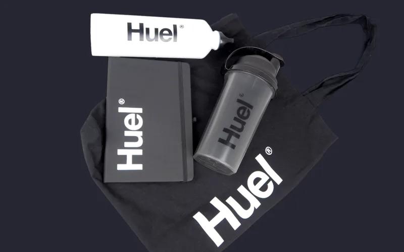 A black tote bag with the Huel logo, a black notebook, a clear shaker bottle, and a white bottle, all featuring the Huel logo.