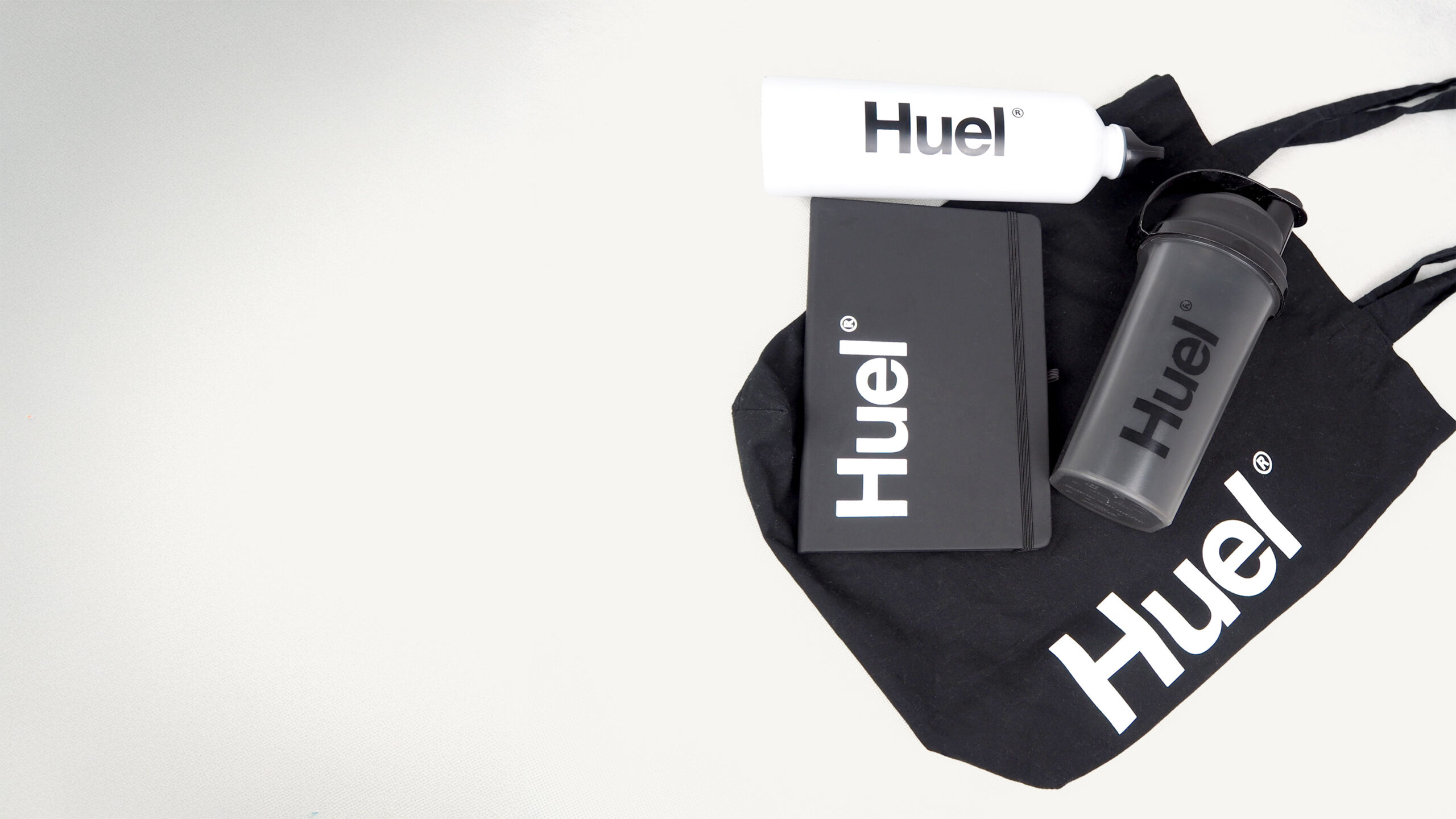A black tote bag, a notebook, a white water bottle, and a dark shaker bottle, all featuring the brand name 