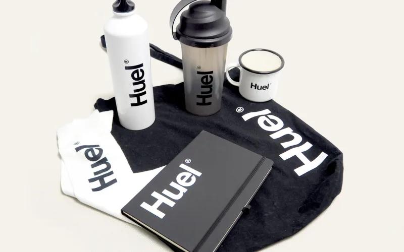 A collection of Huel-branded items, including two water bottles, a shaker, a mug, a notebook, and a black bag.