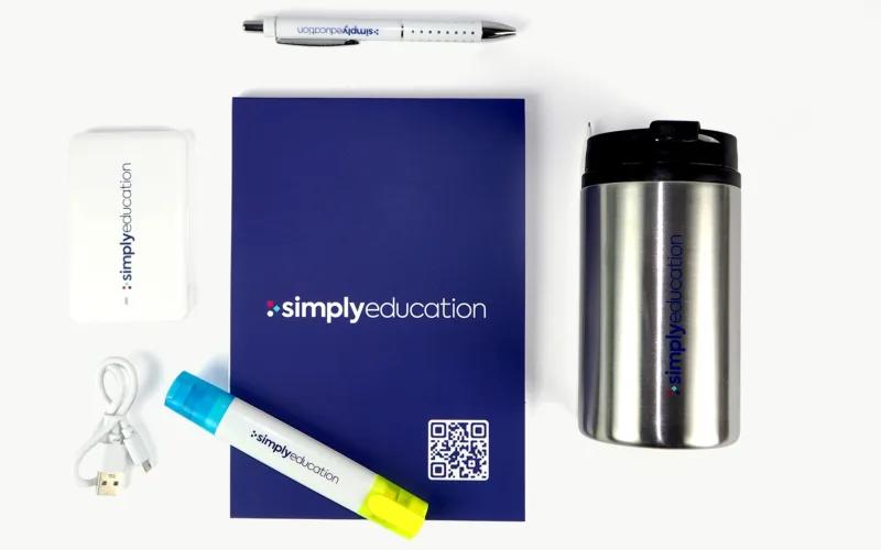 A silver travel mug, a blue-and-yellow highlighter, a pen, a USB cable, a white power bank, and a dark blue notebook with the logo 