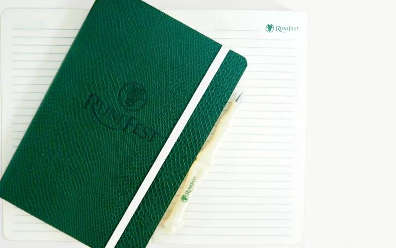 A green notebook with a textured cover and a white elastic band, placed on lined paper with a pen beside it. The notebook has 