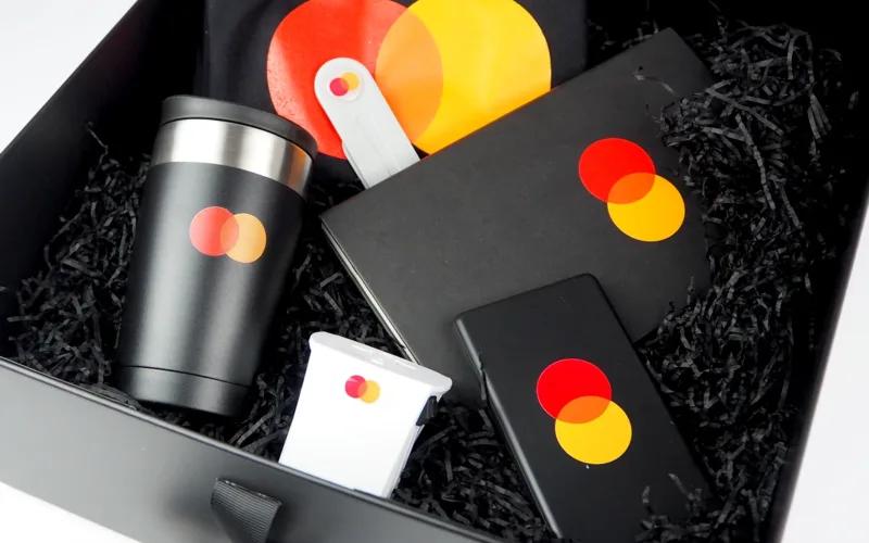 A gift box containing a black travel mug, a notebook, a phone charger, a white device, and a black item, all featuring the Mastercard logo of overlapping red and yellow circles, set against a black shredded paper background.