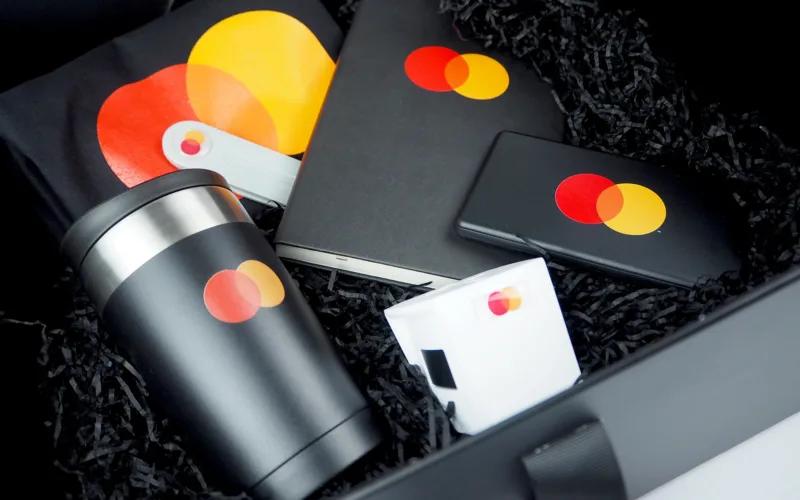 A collection of branded merchandise featuring the Mastercard logo, including a travel mug, notebook, and a mobile charging device, arranged in a black box with shredded paper filling.