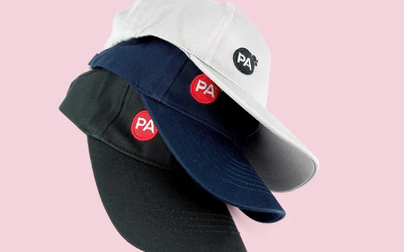 A stack of three baseball caps in white, navy, and black, each featuring a circular logo with 
