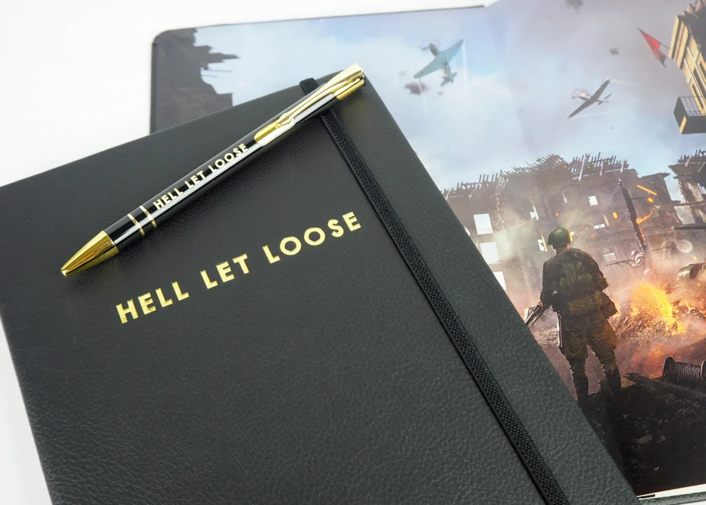 A black notebook with "HELL LET LOOSE" embossed in gold, a matching pen, and an artistic image featuring a soldier and aircraft in a war-torn landscape.