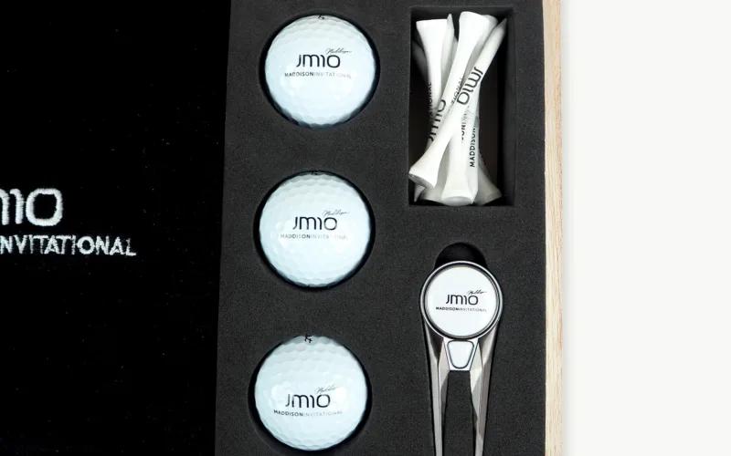 A golf accessory set featuring two golf balls, three golf tees, and a ball marker, all placed in a fitted black foam compartment inside a wooden box. The items are branded with 