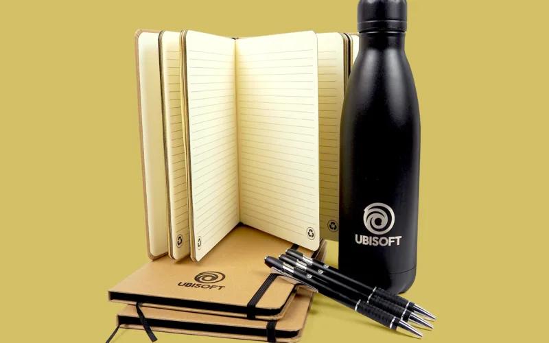 A black water bottle and two notebooks with a logo, accompanied by three pens, all set against a plain background.